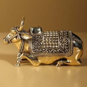 May include: A brass sculpture of a reclining bull with intricate detailing. The bull has a saddle and decorative harness. The sculpture is gold-toned with darker accents, and the text "exotic india" is visible in the upper left corner.