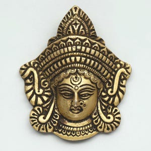May include: A detailed brass sculpture of a deity's face, featuring intricate carvings and a serene expression. The sculpture has a golden hue and is adorned with ornate headwear and jewellery. The image includes the text "exotic india" and "authentic. curated heritage".