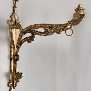Puede incluir: Soporte de pared de latón ornamentado con intrincados adornos y un remate decorativo. El soporte presenta un brazo curvo con un anillo y una placa de montaje vertical. El diseño es detallado y elegante, adecuado para exhibir objetos decorativos.