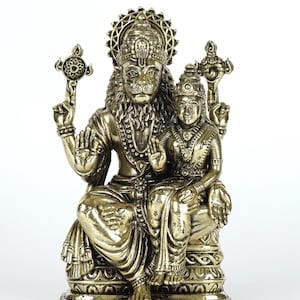 May include: A gold-colored statue depicting a deity with a lion's face and a crown, seated with another figure. The statue features intricate details, including ornate jewelry and a decorative base. The text "exotic india" is visible in the upper left corner.
