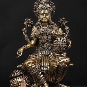 Könnte beinhalten: Eine detaillierte Messingstatue einer sitzenden Gottheit, geschmückt mit aufwendigem Schmuck und Lotusblumen sowie einem dekorativen Topf. Die Figur sitzt auf einer Lotusblütenbasis, mit einem Heiligenschein hinter dem Kopf. Die Statue hat ein Etikett mit der Aufschrift "exotic india".