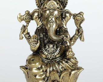 May include: A gold-colored statue of the Hindu deity Ganesha, seated on a lotus flower. The statue features intricate details, including a large head, elephant trunk, and multiple arms holding various objects. The statue has a decorative halo and is from Exotic India.