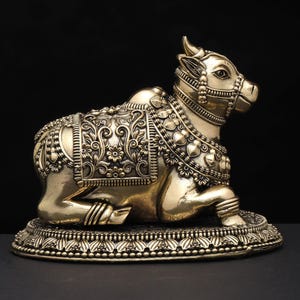 May include: A detailed brass sculpture of a reclining cow, adorned with intricate floral and heart-shaped patterns. The cow is resting on a decorative base, showcasing detailed craftsmanship. The sculpture has a polished finish and is set against a dark background.