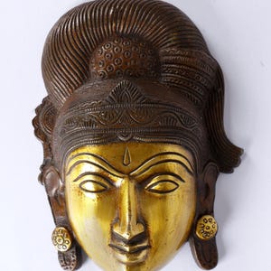 May include: A brass wall hanging depicting a serene face with closed eyes. The face is golden, with detailed features and a decorative headdress. The piece has a vintage aesthetic, with the text "exotic india" at the top.
