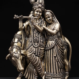 May include: A silver-toned statue depicting a couple, with one figure playing a flute. The figures are adorned in detailed traditional clothing and jewelry. The statue is mounted on a base with a cow figure. The background is black, and the text "exotic india" is visible.