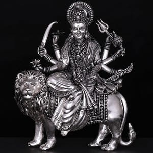 May include: A silver statue of a deity with multiple arms, seated on a lion. The deity wears ornate jewelry and holds various objects, including a sword and trident. The statue has a detailed design and is set against a black background.