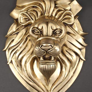 Pode incluir: Escultura de parede dourada em forma de cabeça de leão, com detalhes intrincados da juba e do rosto. A escultura tem uma aparência majestosa, com ênfase nos olhos e na boca aberta. Provavelmente feita de latão ou metal semelhante.