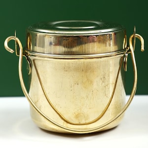 以下が含まれることがあります： 蓋と曲がったハンドルが付いた、金色の金属製容器。容器は円筒形の本体と光沢のある反射面を持っています。ハンドルは、曲がったフックで側面に固定されています。背景は無地の緑色です。
