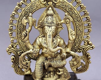 May include: A gold-colored statue of the Hindu deity Ganesha, seated with multiple arms, holding various objects. The statue is set against an ornate, circular backdrop and mounted on a dark wooden base. The statue is approximately 8 inches tall.
