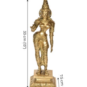 May include: A golden-colored statue of a female figure, standing on a square base. The statue is 13 inches tall and 4 inches wide. The figure is adorned with detailed jewelry and clothing. The base is 3 inches tall. The statue is from Exotic India.