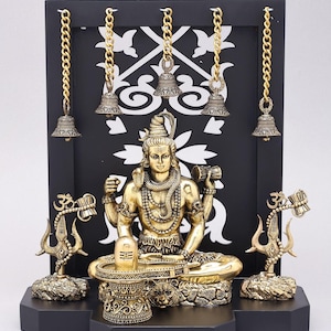 May include: A gold-colored statue of a seated deity with multiple arms, adorned with necklaces, and a serene expression. The statue is set against a black backdrop with hanging bells and decorative elements. The base is black.