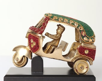 May include: A gold-coloured, three-wheeled auto-rickshaw figurine with red and green beaded accents. The figurine features a driver and passenger seat, mounted on a black base. The top of the rickshaw is green with gold and red accents.