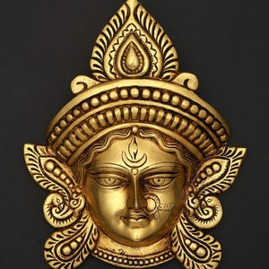 May include: A golden brass sculpture of a deity's face, with detailed features. The face has a calm expression, with a nose ring and an elaborate headdress. The sculpture is set against a dark background, accentuating its ornate design.