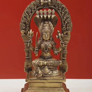May include: A brass statue of a seated deity, adorned with a crown and multiple cobra heads. The figure holds a sword and a trident, set against a red backdrop. The base of the statue is rectangular, with the text "exotic india" at the top.