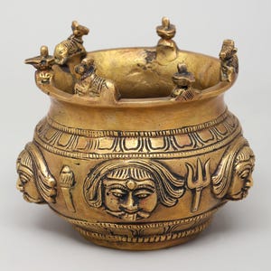 May include: A gold-coloured, antique-style brass pot with intricate carvings. The pot features detailed faces and figures around the exterior and small statues around the rim. The pot has a wide opening and a textured surface. The text "exotic india" is visible.