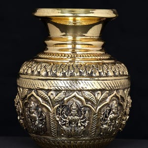 May include: A gold-colored, ornate metal vase with a wide base and a flared rim. The vase features detailed relief carvings of figures and floral patterns. The text "exotic india" is visible at the top.