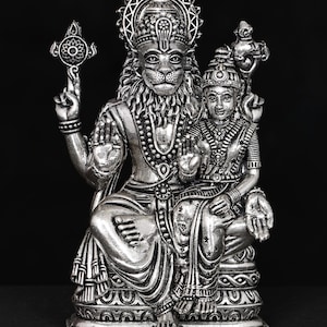 May include: A detailed silver statue of a deity with a lion-like face and a crown, seated with a female figure. The statue features intricate carvings, necklaces, and armbands. The background is black, and the statue is labeled "exotic india".