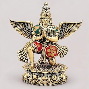 Può includere: Statua color oro di una figura in ginocchio con grandi ali dettagliate. La figura indossa una corona, abiti verdi e rossi e ha le mani giunte in posizione di preghiera. La statua è su una base decorativa.