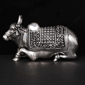 May include: A silver-colored sculpture of a seated bull with detailed decorative saddle and ornate bridle. The sculpture is set against a black background. The text "exotic india authentic curated heritage" is in the top left corner.