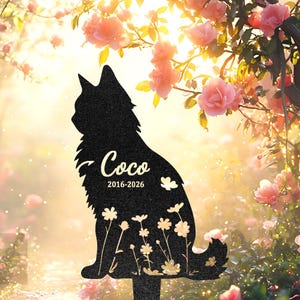 May include: Black metal cat silhouette garden stake with the name "Coco" and the years 2016-2026. The cat is surrounded by a floral design and is set against a backdrop of pink flowers and sunlight.