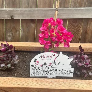 May include: A white metal garden stake featuring a cutout silhouette of a sleeping cat, the name Micro, the years 2012-2026, and a decorative pattern of black flowers, stems, and butterflies, positioned upright in a soil-filled wooden planter box.