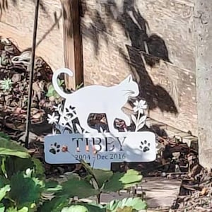 May include: A white metal garden stake featuring a silhouette of a cat walking among flowers. Below the cat, a rectangular plaque displays the name TIBEY, the years 2004 - Dec 2016, and two paw print icons, one on each side of the text.