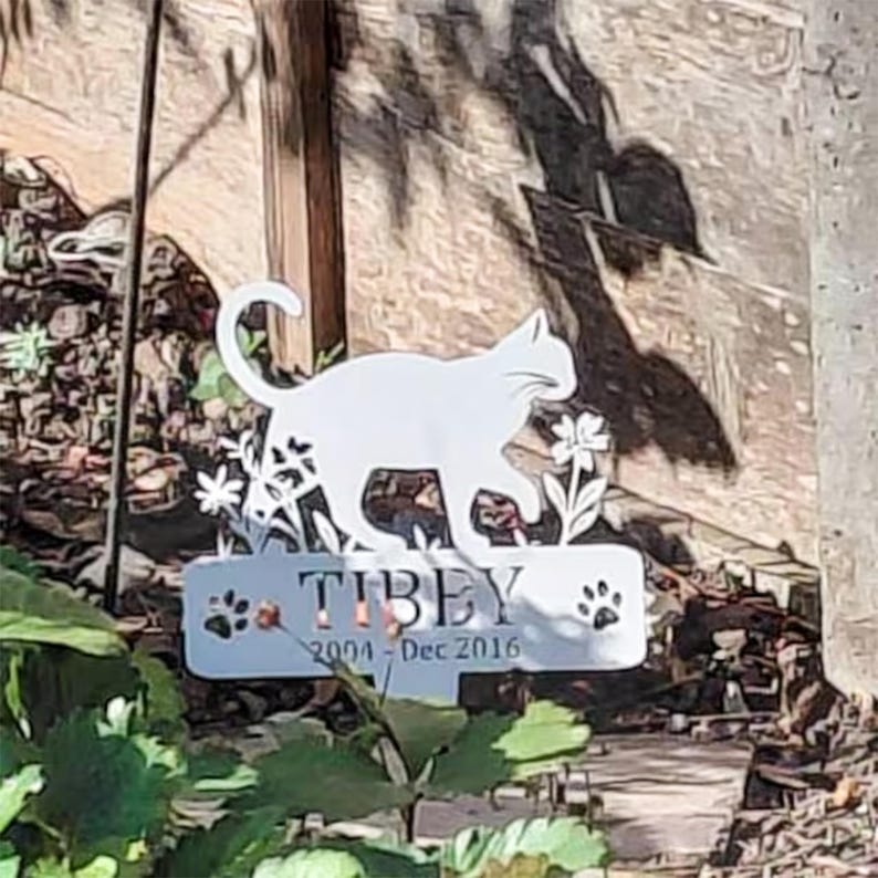 May include: A white metal garden stake featuring a silhouette of a cat walking among flowers. Below the cat, a rectangular plaque displays the name TIBEY, the years 2004 - Dec 2016, and two paw print icons, one on each side of the text.