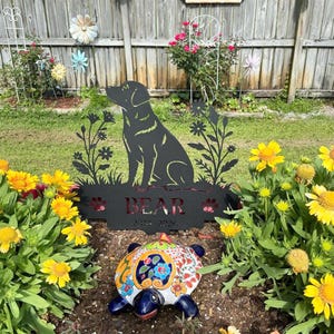 May include: A black metal garden sign featuring a silhouette of a sitting dog surrounded by floral cutouts. The sign includes the name BEAR and the years 2014-2024 cut into the metal. A colorful ceramic turtle figurine sits in front of the sign.