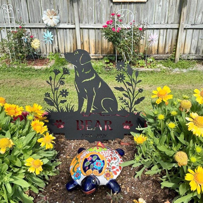 May include: A black metal garden sign featuring a silhouette of a sitting dog surrounded by floral cutouts. The sign includes the name BEAR and the years 2014-2024 cut into the metal. A colorful ceramic turtle figurine sits in front of the sign.