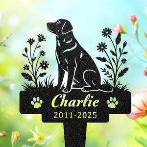 May include: A black metal garden stake featuring a silhouette of a sitting dog flanked by floral stems. The central plaque displays the name Charlie in a script font, flanked by two paw prints, with the years 2011-2025 printed below in yellow.