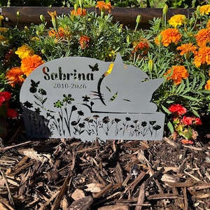 May include: A grey metal garden memorial stake featuring a cutout silhouette of a sleeping cat, floral patterns, and a butterfly. The text "Sabrina" and "2010-2026" are laser-cut into the surface. The design includes a pointed base for insertion.