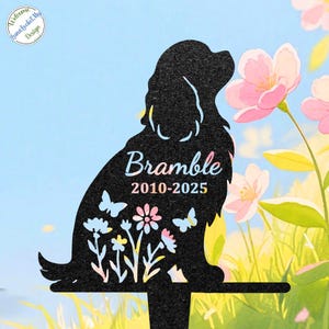 May include: Black metal garden stake in the shape of a dog, with the name "Bramble" and the years "2010-2025" in script. The dog silhouette features cut-out flowers and butterflies. The background shows pink flowers and a blue sky.