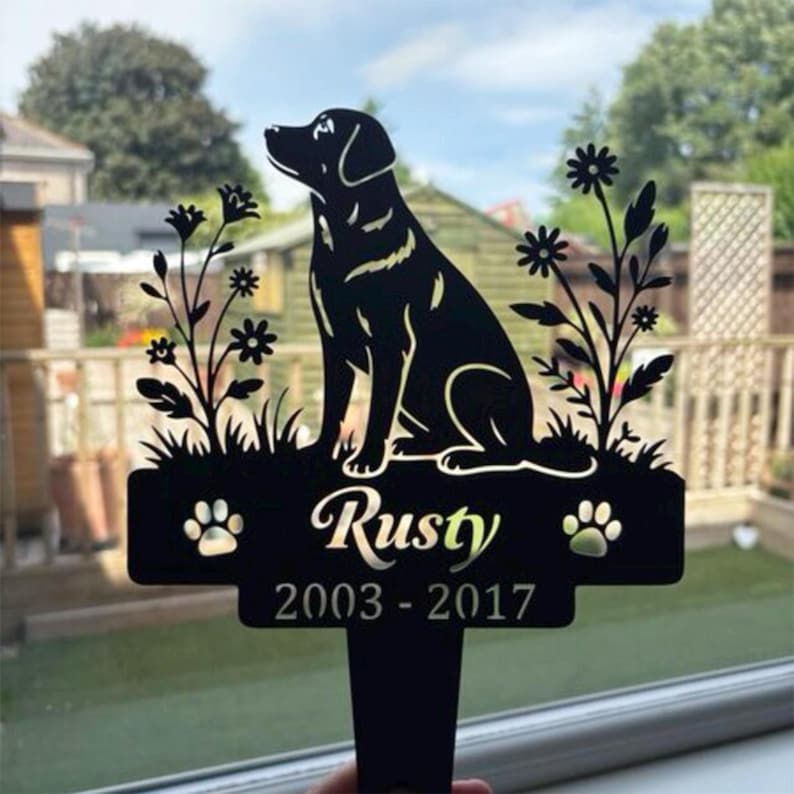 May include: A black metal garden stake featuring a silhouette of a sitting dog surrounded by flowers. The base includes the name Rusty in script font, two paw prints, and the years 2003-2017 engraved in a clean, sans-serif typeface.