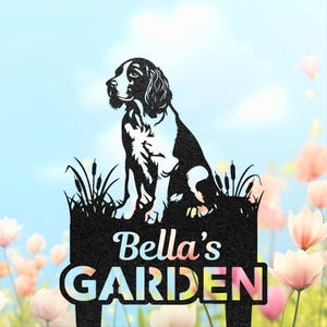 May include: Black metal garden sign with a dog silhouette sitting in front of a sign that reads "Bella's GARDEN". The dog is black and white, and the sign is surrounded by grass and reeds. The text is in a colourful font.