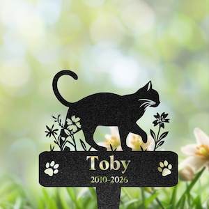 May include: A black metal memorial garden stake featuring a silhouette of a cat walking among flowers. The rectangular base displays the name Toby and the years 2010-2026, flanked by two paw prints on either side of the text in a clean font.