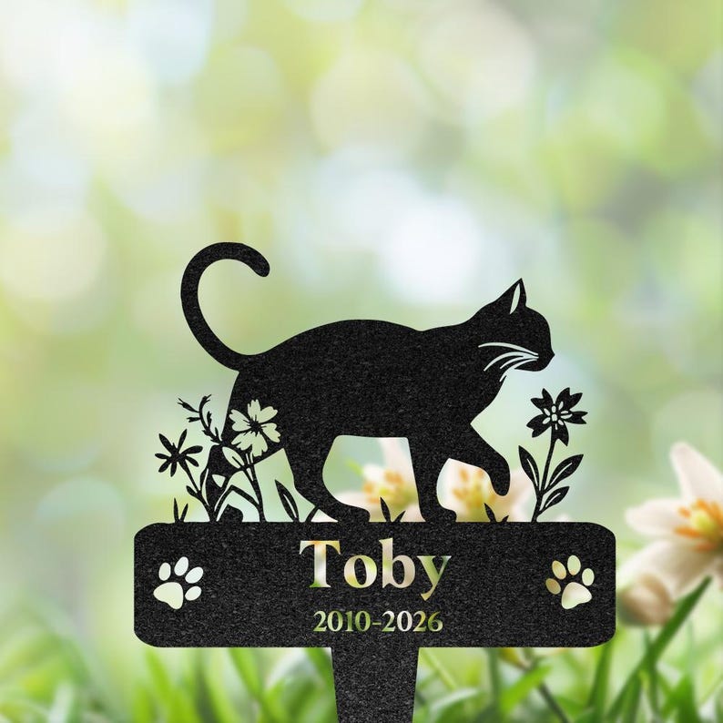 May include: A black metal memorial garden stake featuring a silhouette of a cat walking among flowers. The rectangular base displays the name Toby and the years 2010-2026, flanked by two paw prints on either side of the text in a clean font.