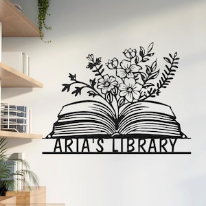 May include: Black metal wall art featuring an open book with floral accents. The book's pages are detailed, and flowers and leaves sprout from the center. The text "ARIA'S LIBRARY" is displayed below the book.