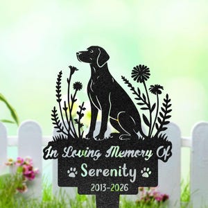 May include: Black metal garden stake featuring a silhouette of a dog sitting among stylized flowers. The stake includes the text "In Loving Memory Of Serenity" with paw prints and the years 2013-2026.
