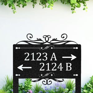 Könnte beinhalten: Schwarzes Metall-Adressschild mit dekorativen Schnörkeln. Das Schild zeigt die Nummern 2123 A mit einem Pfeil nach rechts und 2124 B mit einem Pfeil nach links. Das Schild ist von Grün umgeben.