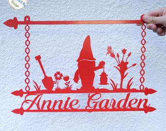 May include: Red metal garden sign with a gnome holding a lantern, a shovel, flowers, and a butterfly. The sign reads "Annie Garden" in cursive. The sign is suspended by chains and has decorative arrow-shaped ends.