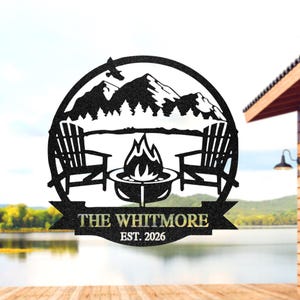 Op de afbeelding: Zwarte metalen wandkunst met een berglandschap, twee Adirondack stoelen, een vuurplaats en de tekst "THE WHITMORE EST. 2026". Het ontwerp is geplaatst in een cirkelvormig frame, ideaal voor buiten- of hutdecoratie.