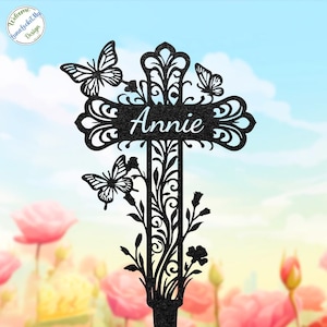 May include: Black metal garden stake featuring a cross design with butterflies and floral accents. The name "Annie" is displayed in white script. The stake is designed for outdoor use and adds a decorative touch to gardens.