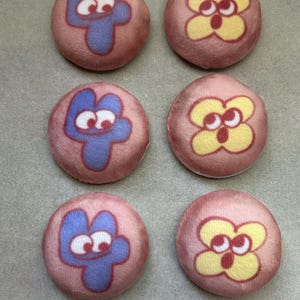 May include: Six round, pink fabric buttons. Three buttons feature a blue number four with white eyes. The other three buttons have a yellow flower design with red outlines and white eyes. The buttons are arranged in two rows of three.