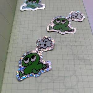 May include: Three holographic stickers featuring a green cartoon character with large eyes and a thought bubble. The thought bubble contains a musical note. The stickers are on a grid-lined paper.