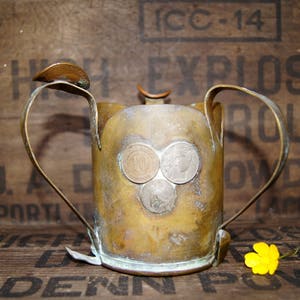 Puede incluir: Una jarra de latón antigua con dos asas curvas y tres monedas adheridas al frente. La jarra tiene un aspecto deslustrado y envejecido. El fondo es una caja de madera con texto descolorido.