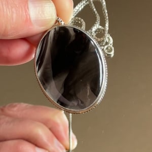 May include: A silver necklace with a large, oval, black pendant. The pendant has a shiny surface and is set within a decorative silver frame. The chain is fine and the image shows the pendant held up, with a reflection of the pendant below.
