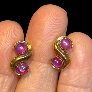 May include: A pair of gold-toned earrings, each featuring a stylized S-shape design. Each earring has two round, deep pink gemstones. The earrings are displayed against a dark background.