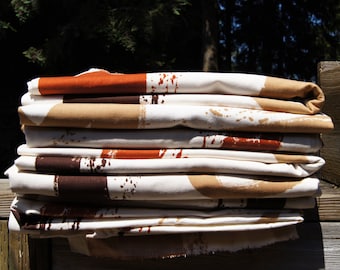 May include: A stack of folded fabric with a white base, featuring brown, tan, and rust-colored abstract designs. The fabric has a splattered paint effect, adding a unique touch to the textile.