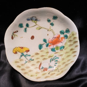 May include: A small, off-white ceramic plate with a scalloped edge. The plate is decorated with hand-painted flowers in orange, yellow, and blue, along with green leaves and stems. A rooster is also painted on the plate.