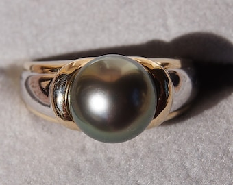 May include: A gold and silver ring featuring a large, dark gray pearl. The ring has a wide band with gold accents around the pearl. The pearl is round and has a lustrous surface.
